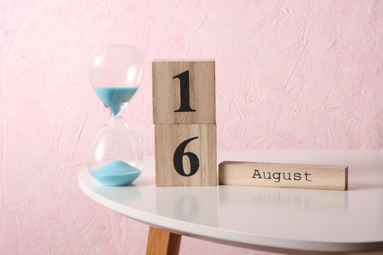 August 16 wooden block calendar and hourglass on table, pink concrete background