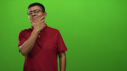 Fototapeta premium Young man with hand on chin posing in studio green backdrop wearing red polo and glasses; thoughtful reflection.
