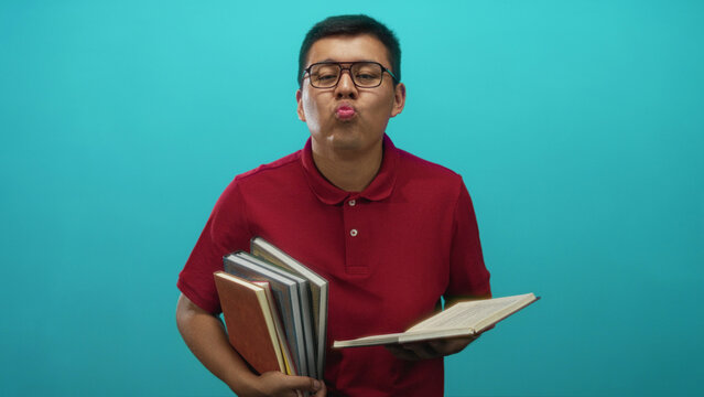 Hispanic man wearing glasses and a red polo shirt holding stacked books and an open book while puckering his lips in a turquoise studio; playful study focus.