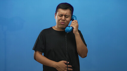 Young man holding blue handset to ear in studio with closed eyes and pained expression; discomfort awkward call.