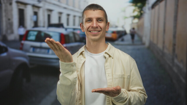 Man gesturing on street with hands raised in an urban outdoor setting, wearing casual clothes and smiling at camera with blurred cars in background.