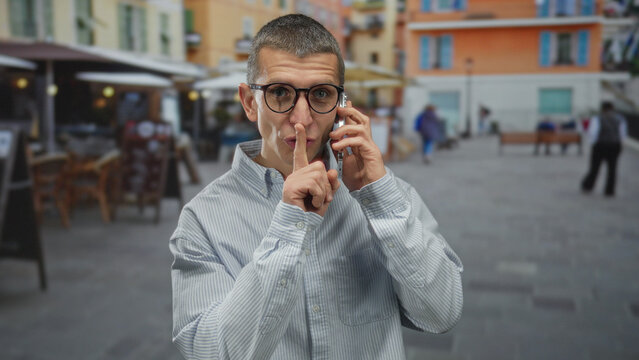 Man talking on smartphone outdoors on a busy street terrace, surrounded by colorful buildings and restaurant seating, wearing a striped shirt and glasses.