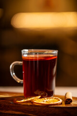 Glass of steaming mulled wine with dried orange slices and a cinnamon stick on a wooden board against a blurred warm background.