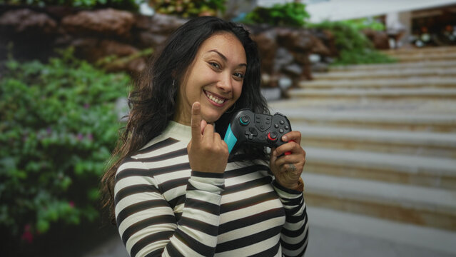 Woman holds a game controller with both hands and smiles while standing on building steps near greenery; joyful play.