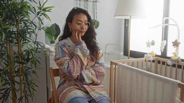 Woman seated by a crib in a building with hand on cheek and colorful cardigan, wearing glasses; motherhood concern. - Powered by Adobe