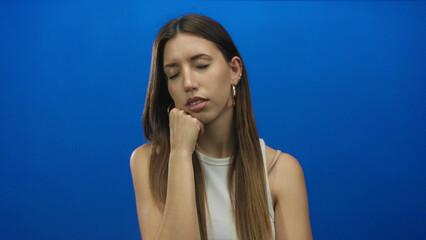 Woman with hand supporting chin and eyes closed in a vivid blue studio setting wearing a white tank...
