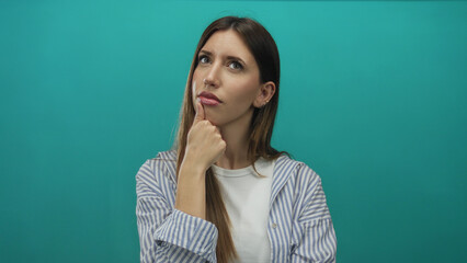 Obraz premium Young hispanic woman wearing striped shirt with hand on chin in teal studio scene; contemplation doubt.
