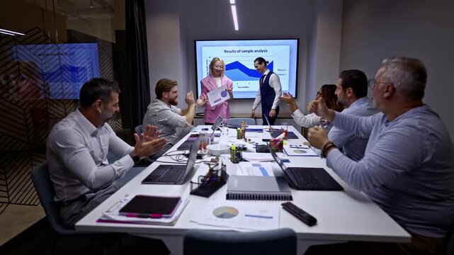 Female presenter concludes a successful business presentation, receiving applause and recognition from her diverse team of coworkers in a contemporary meeting room. They discuss results and analysis.