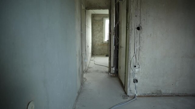 Unfinished apartment corridor with wiring, pipes, and window. Exposed cables, bare plaster, concrete floor, dust, and cool light. Shallow depth of field shows rough texture.