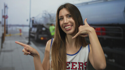 Woman cheerleader in uniform makes a call gesture with her right hand and points her left finger toward a fuel tanker on an airport tarmac; joy.