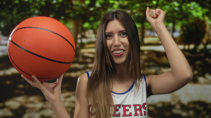 Woman in uniform holds bright orange basketball with fingertips in bright sunlit green park; confidence energy vitality fun.