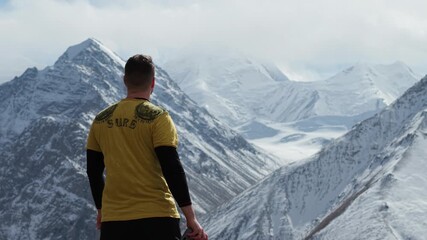Adventurer in yellow shirt stands on rocky terrain, gazing at breathtaking snow-capped mountains, clouds drift above, camera pans slowly to capture the expansive landscape and serene atmosphere