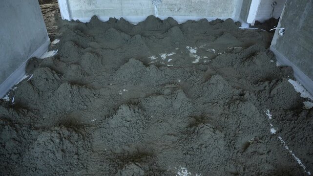 Wet screed mounds on subfloor during interior renovation work. Irregular deposits, wiring loops, rough wall edges, ridge patterns, tool marks, boot prints, cool daylight, low contrast.