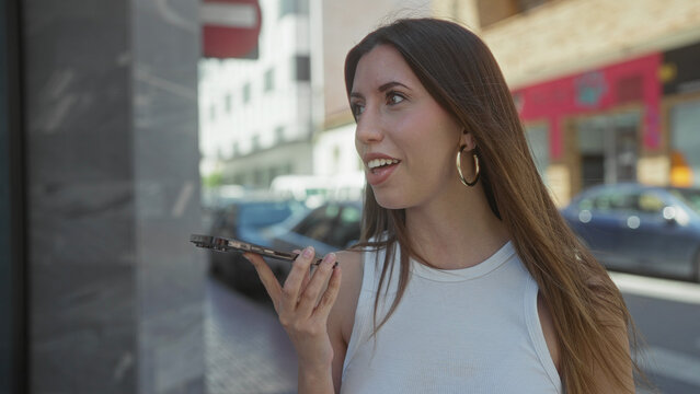 Woman holds smartphone to lips and leans forward while smiling and speaking on a bustling city street; confidence.