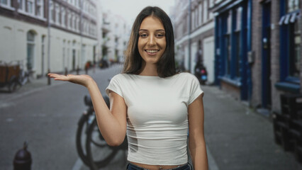Fototapeta premium Woman smiling with palm up presenting gesture on street with white shirt and jeans, hand extended at waist level; friendly invitation.