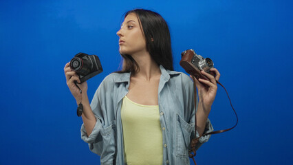 Young hispanic woman holding two cameras in blue studio comparing digital and vintage equipment while looking sideways; creative contemplation.