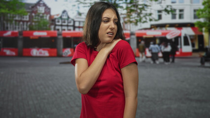 Woman in red shirt with hand on shoulder wincing while standing on city street near a red bus and cafes; pain recovery.