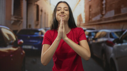 Young hispanic woman with hands pressed together in prayer on a sunlit street between parked cars; serenity gratitude.