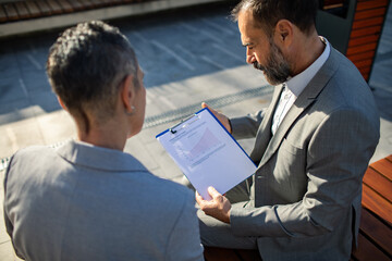 Adult coworkers reviewing financial report with focused expressions on outdoor bench