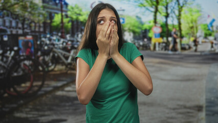 Fototapeta premium Woman covers mouth with hands on busy city street beside parked bicycles, wearing green tee; surprise.