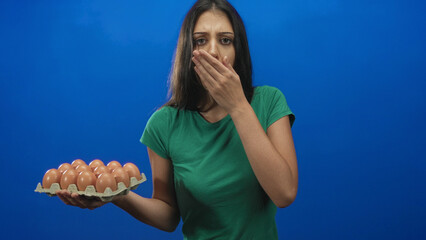 Young hispanic woman holding a carton of eggs in one hand and covering her mouth with the other in...
