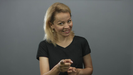 Young blonde woman smiling and pointing finger at camera in gray studio setting; confidence empowerment engagement.