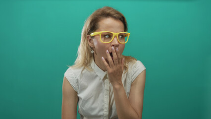 Woman wearing yellow eyeglasses and white blouse raises her hand to mouth while yawning in a teal...