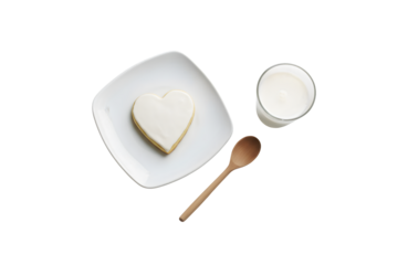 a heart-shaped white cookie sitting on a white square plate next to a glass of milk
