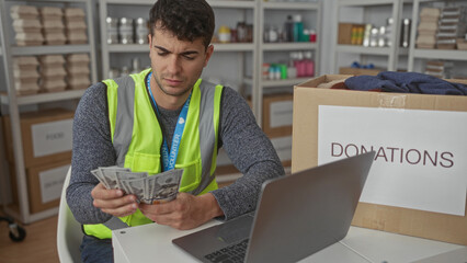 Man held cash while counting records in a warehouse by a hispanic volunteer beside a donation box near a laptop tracking money.