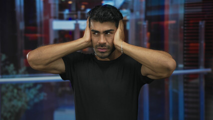 Man pressing hands over ears in hotel building with a tense expression and visible muscle definition; noise annoyance discomfort stress.