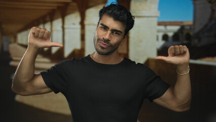Young man in black shirt points thumbs to chest at historic stone building under sunlight; confidence.