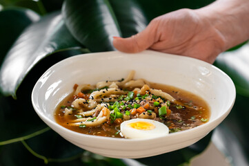 A hand holds a white bowl of hot noodle soup with udon noodles, broth, a boiled egg, vegetables, and black sesame seeds, set against a backdrop of large green leaves.