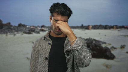 Man pinches nose in street scene by sandy beach and rocky ocean shore under cloudy sky in soft...