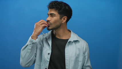 Young hispanic man wearing denim shirt makes pinch gesture with fingers near lips in blue studio; satisfaction joy pleasure delight.