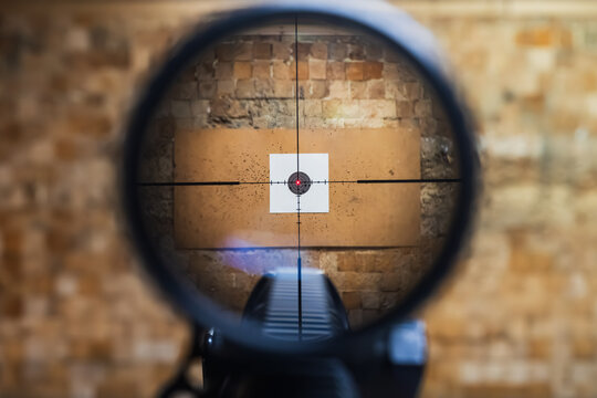 View through a sniper rifle scope aiming at a paper target with bullet holes in an indoor shooting range
