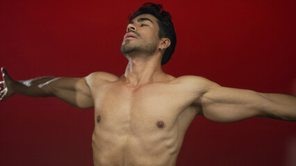 Obraz premium Man with bare chest and outstretched arms in a red studio setting showing muscular torso and shoulders; serenity.