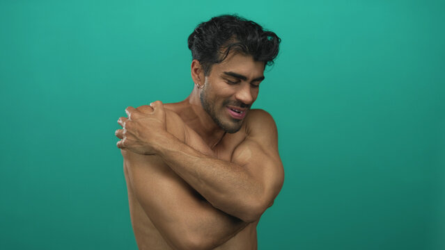 Shirtless man holds his bare shoulder and wraps arms around his chest against green studio wall; serenity.