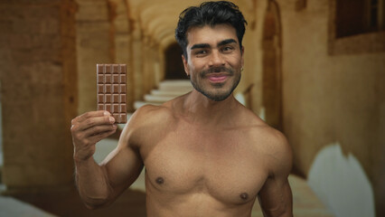 Shirtless hispanic man holds chocolate bar and points finger to object in university corridor; temptation joy.