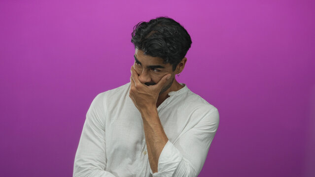 Young man in white shirt covers mouth with hand under purple studio lighting while tilting head downward; hesitation.