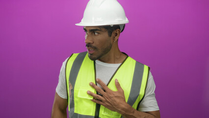 Hispanic engineer man wearing white hardhat and reflective vest touches his chest in a studio; concern safety planning.