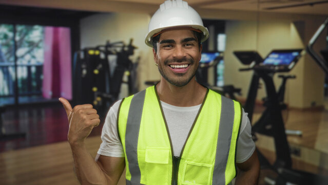 Man wearing hardhat and neon vest smiles and gives thumbs up gesture in gym building; confidence safety.