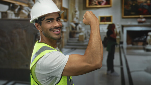 Young hispanic engineer man flexes bicep while wearing hardhat and reflective vest inside building; confidence strength.