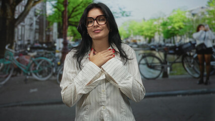 Fototapeta premium Woman clutching chest and smiling beside parked bicycles by a city canal outdoors wearing glasses and striped shirt; gratitude.