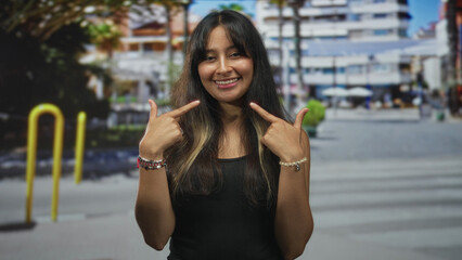 Woman pointing fingers to her cheeks and smiling on a busy urban street with blurred buildings and yellow bollards; joy confidence optimism.