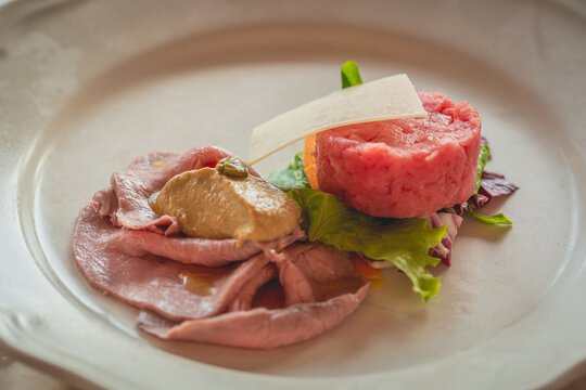 Vitello tonnato and fassona beaf tartar garnished with a Parmesan shaving and fresh salad prepared in the style of the Piedmont region of Italy on a white dish