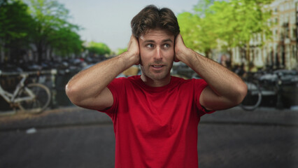 Fototapeta premium Man in red shirt covers ears with hands on amsterdam canal street lined with bicycles and riverside buildings, showing discomfort and raised eyebrows; frustration.