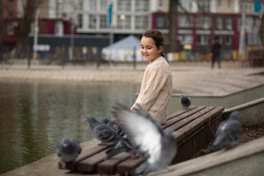 Cheerful young girl in a cozy knit sweater sits on bench by a pond, smiling as pigeons gather around her in a peaceful urban park setting