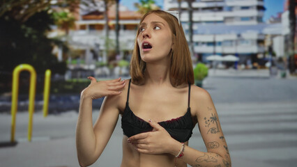 Young blonde woman fans face with hand and shields eyes under sunglasses on busy urban street; heat discomfort.