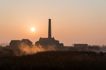 A sunrise over a field with a large brick building in the background. The sky is orange and the sun is rising.