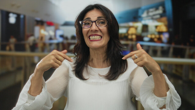 Smiling woman waves hand in bustling mall center with blurred shoppers and railings behind her; friendly joy.